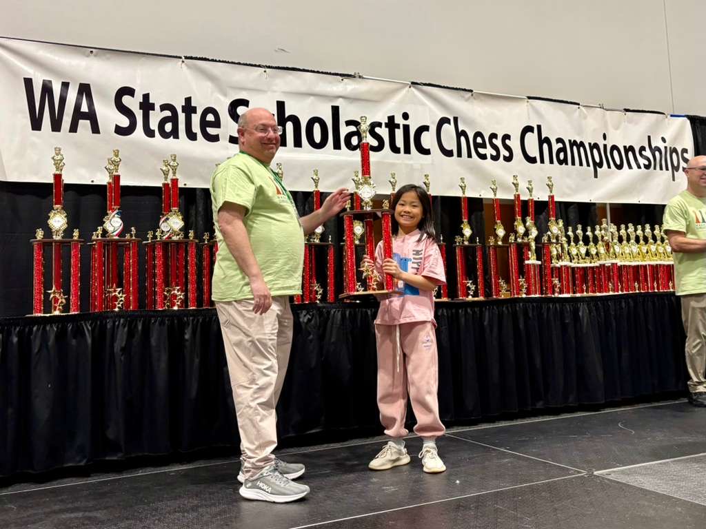 A young girl stands with a tournament organizer holding a large trophy in front of a banner reading 'WA State Scholastic Chess Championships,' with rows of trophies displayed behind them